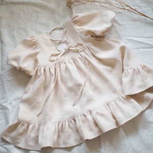 May include: A light beige short-sleeved top featuring puff sleeves, a ruffled hem, and a lace-up back closure with matching fabric ties. The garment has a gathered bodice and a loose, flowing silhouette, showcasing a textured, woven fabric finish.