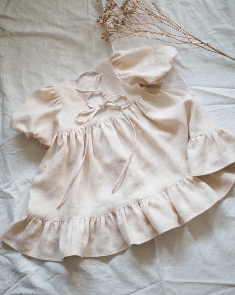 May include: A light beige short-sleeved top featuring puff sleeves, a ruffled hem, and a lace-up back closure with matching fabric ties. The garment has a gathered bodice and a loose, flowing silhouette, showcasing a textured, woven fabric finish.