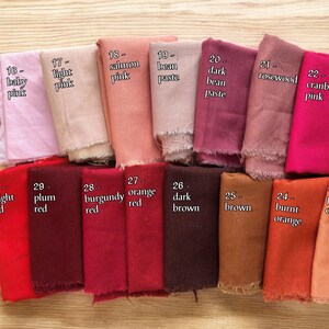 Op de afbeelding: Een verzameling van 14 stofstalen in verschillende tinten roze, rood, oranje en bruin. Elk staal is gelabeld met een nummer en een kleurnaam, zoals "baby roze", "pruim rood" en "gebrand oranje".