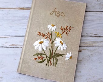 May include: A beige linen notebook with the name "Asa" embroidered on the front cover. The notebook features an embroidered design of white daisies with green stems and leaves.
