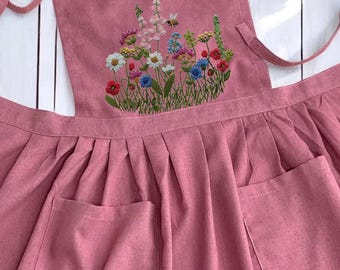 May include: A pink linen apron with a floral embroidery design featuring a variety of flowers and a bee. The apron has two large pockets and a tie at the neck.