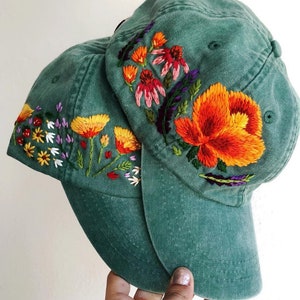 Puede incluir: Dos gorras de béisbol verdes con diseños florales bordados. Las gorras presentan una variedad de flores coloridas, incluyendo naranja, amarillo, rojo, rosa y morado.