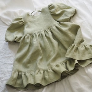 May include: A sage green dress featuring short puff sleeves with gathered cuffs, a round neckline with a tie closure at the back, and a gathered bodice that transitions into a flared skirt with a wide, ruffled hemline along the bottom edge.