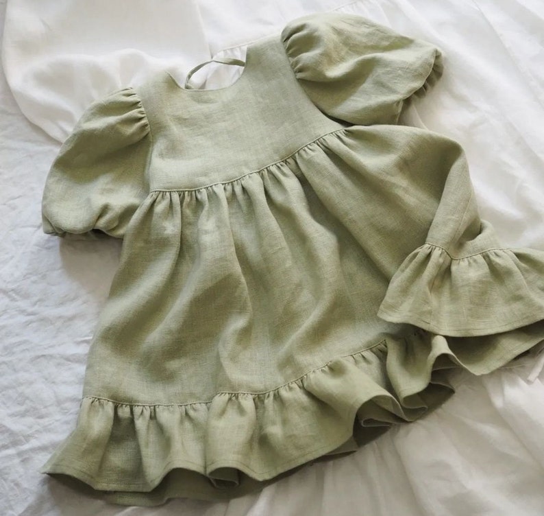 May include: A sage green dress featuring short puff sleeves with gathered cuffs, a round neckline with a tie closure at the back, and a gathered bodice that transitions into a flared skirt with a wide, ruffled hemline along the bottom edge.