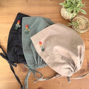 Puede incluir: Tres sombreros de tela con lazos ajustables. Los sombreros son de color negro, verde y beige. Cada sombrero tiene un pequeño diseño de flor bordada con hojas verdes.