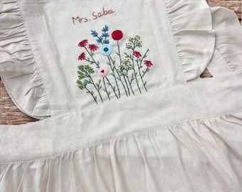 May include: A white linen apron with a ruffled neckline and embroidered flowers and the text "Mrs. Saba".