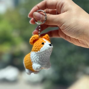 May include: A handmade orange and white crochet corgi dog keychain. The corgi has black eyes and a silver keychain ring. The dog is held up by a hand against a blurred background.