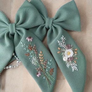 Puede incluir: Dos lazos para el pelo de tela verde salvia con dise&ntilde;os florales bordados. Un lazo presenta un ramo de flores blancas, rosas y marrones, mientras que el otro tiene una sola flor blanca con detalles rosas y marrones.