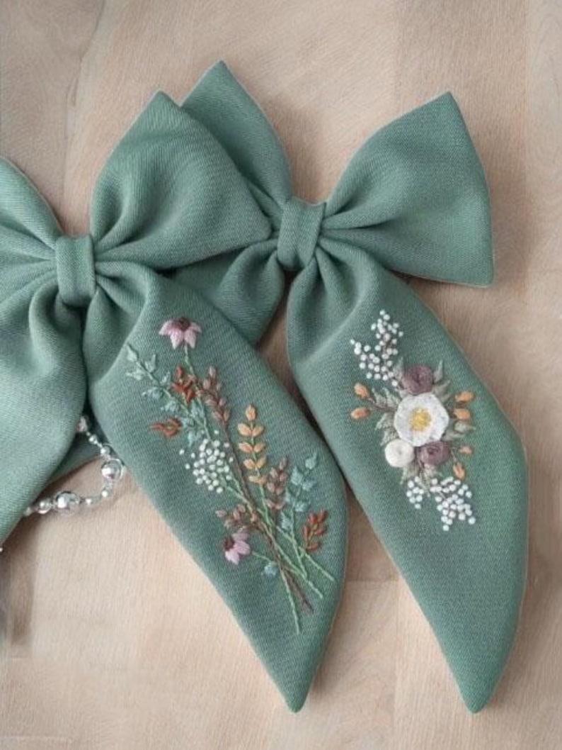 Puede incluir: Dos lazos para el pelo de tela verde salvia con dise&ntilde;os florales bordados. Un lazo presenta un ramo de flores blancas, rosas y marrones, mientras que el otro tiene una sola flor blanca con detalles rosas y marrones.