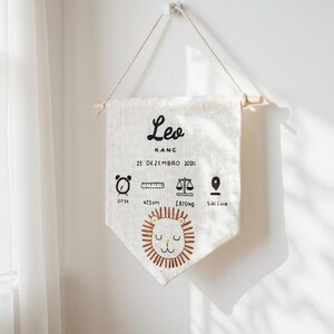 May include: A cream-colored banner with the name "Leo" in black script, along with birth details and a lion illustration. The banner includes measurements of 47.5 cm and a weight of 2.870 kg. It hangs from a wooden dowel and a white wall.