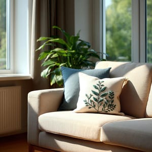 Peut inclure: Un canapé beige avec deux coussins décoratifs. L'un est bleu clair, l'autre est crème avec un motif de feuilles vertes brodées. Une grande plante verte est visible en arrière-plan près d'une fenêtre.