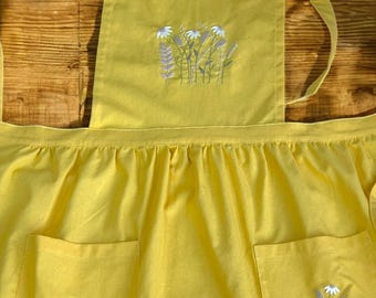 May include: A yellow apron with two pockets, each embroidered with a cluster of white flowers and green leaves. The apron has a single strap that goes over the head and ties at the neck.