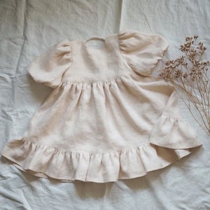 May include: A light beige dress with short puff sleeves, a gathered waistline, and a ruffled hem. The garment features a round neckline with a small back closure detail. The fabric has a visible woven texture, and the design is simple and plain.