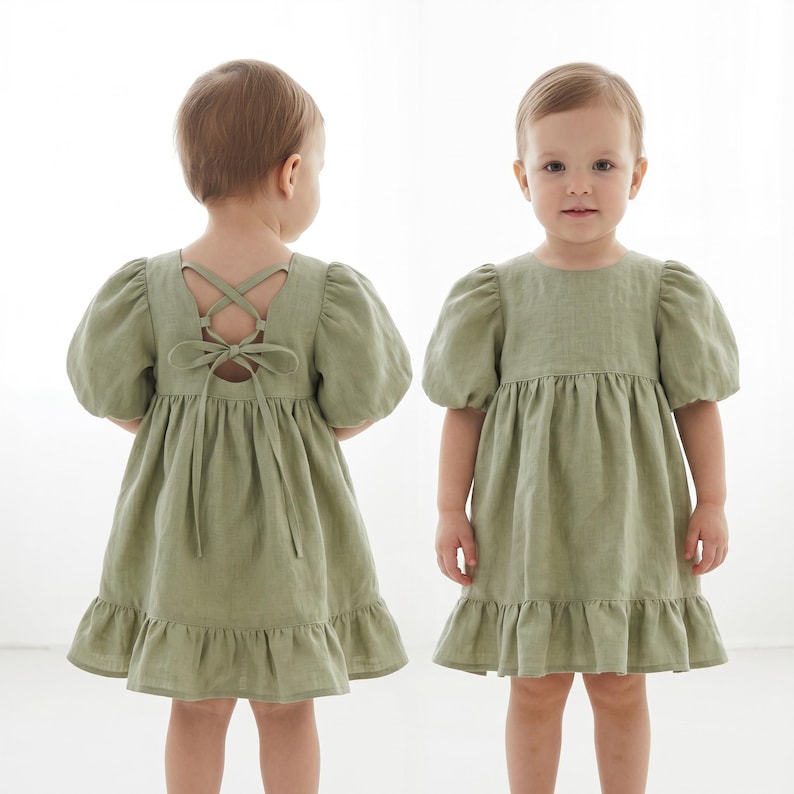 May include: A sage green dress with puff sleeves, a round neckline, and a ruffled hem. The back features a cutout with crisscross tie-up detailing. The garment is shown from both the front and back views to display the design and construction.