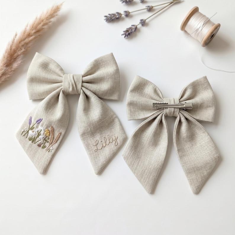 May include: Two beige linen hair bows. One bow features embroidered wildflowers and wheat on the left tail and the name Lilly embroidered on the right tail. The second bow shows a silver metal alligator clip attached to the back centre.