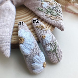 May include: Two beige fabric hair clips with embroidered white flowers and green leaves. The clips are labeled #1 and #2.