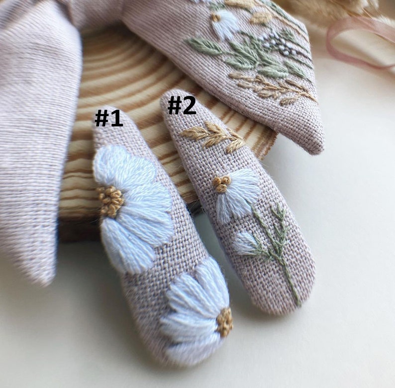 May include: Two beige fabric hair clips with embroidered white flowers and green leaves. The clips are labeled #1 and #2.
