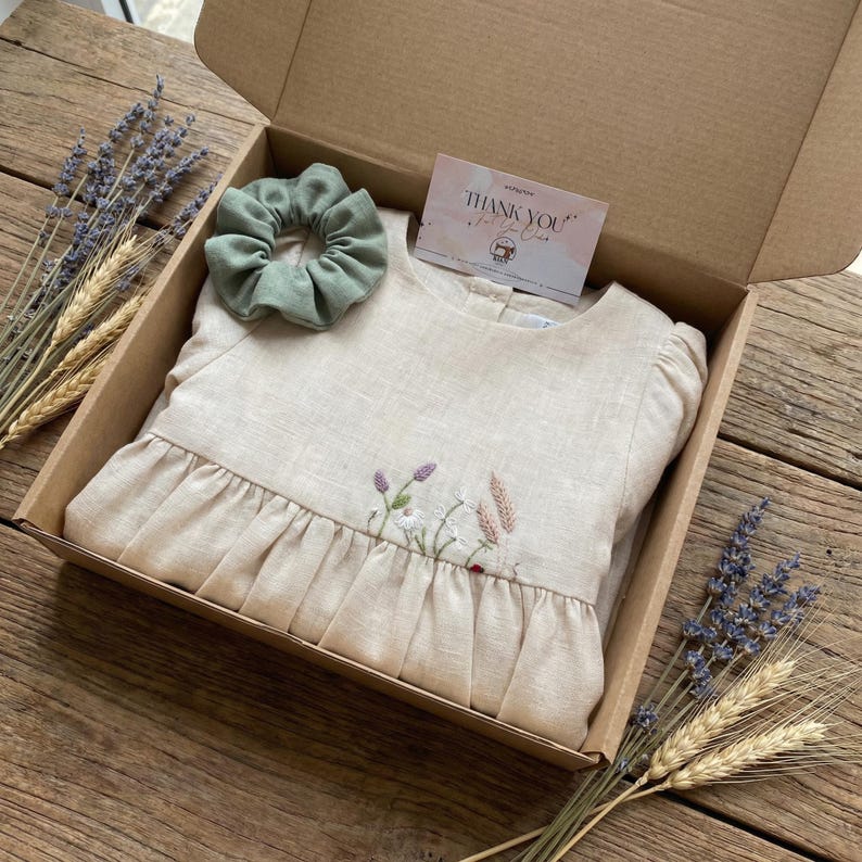 May include: A beige linen dress with floral embroidery, a sage green scrunchie, and a thank you card inside a cardboard box. The dress features a gathered waist and long sleeves. Dried lavender and wheat stalks are placed around the box.