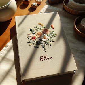 Op de afbeelding: Een beige linnen dagboek met een geborduurd bloemenmotief met roze, witte en rode bloemen. De naam "Elly" is in rood onder de bloemen geborduurd.