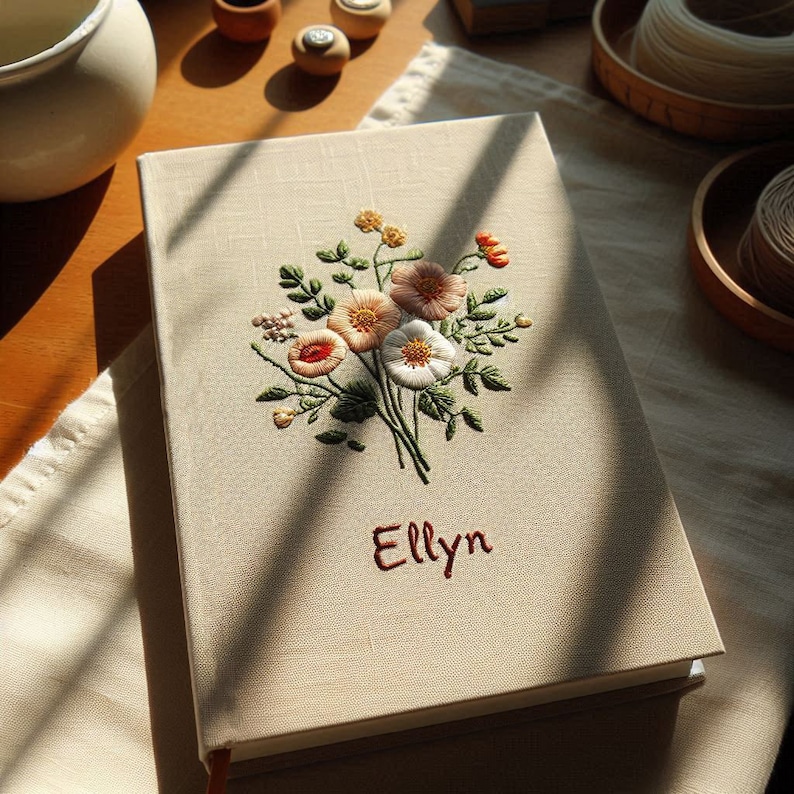 Op de afbeelding: Een beige linnen dagboek met een geborduurd bloemenmotief met roze, witte en rode bloemen. De naam "Elly" is in rood onder de bloemen geborduurd.