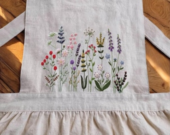 May include: A beige linen apron with a floral embroidery design. The embroidery features a variety of flowers in shades of pink, purple, red, and green.
