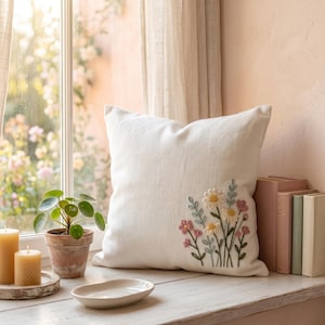 Könnte beinhalten: Ein quadratisches weißes Zierkissen mit einem gestickten Blumenmuster in der unteren rechten Ecke. Die Stickerei zeigt Gänseblümchen mit gelben Mitten, kleine rosa Blüten sowie grüne Stängel und Blätter. Das Kissen hat eine strukturierte Oberfläche.