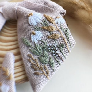May include: A beige linen fabric with embroidered white flowers and brown stems. The fabric has a textured surface and is perfect for crafting.