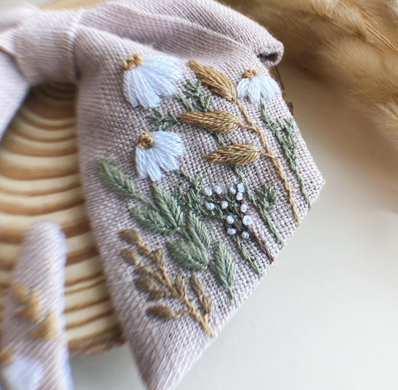 May include: A beige linen fabric with embroidered white flowers and brown stems. The fabric has a textured surface and is perfect for crafting.