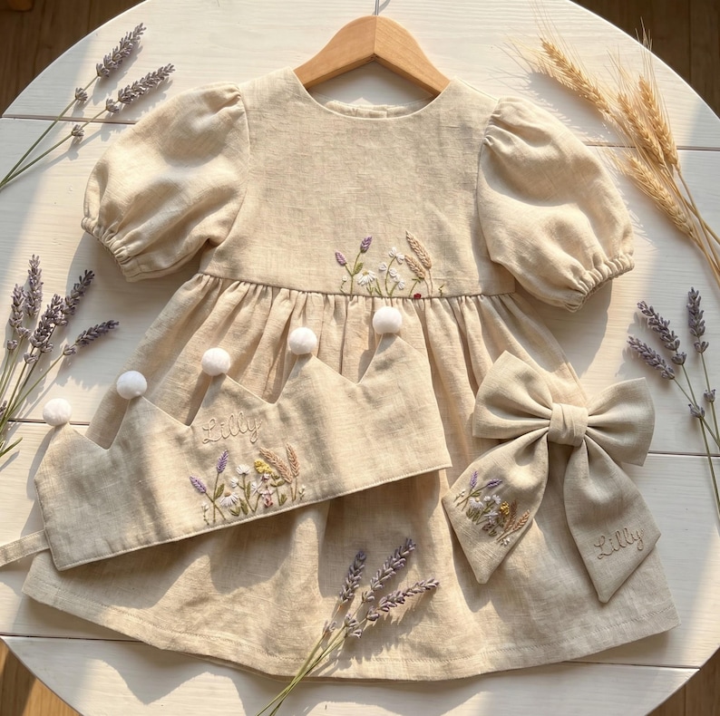 May include: A beige dress with puff sleeves, a matching crown with white pom-poms, and a hair bow. The dress, crown, and bow feature embroidered floral designs and wheat stalks. The name Lilly is embroidered on the crown and the hair bow.