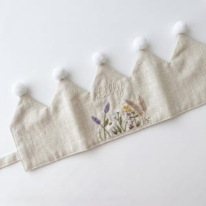 May include: A beige fabric crown featuring five points topped with white pom-poms. The centre point is embroidered with the name Lilly in white script above a detailed floral arrangement of lavender, daisies, and wheat stalks in natural tones.