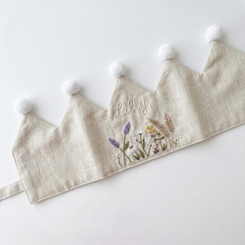 May include: A beige fabric crown featuring five points topped with white pom-poms. The centre point is embroidered with the name Lilly in white script above a detailed floral arrangement of lavender, daisies, and wheat stalks in natural tones.