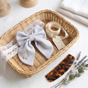 Peut inclure: Un nœud pour cheveux en tissu gris clair avec une oie blanche brodée aux pattes orange, fixé sur un bandeau en dentelle blanche. Il comprend un ruban crème avec une étiquette "HANDMADE & LOVED & LINEN & GOOSE" et un peigne en écaille.