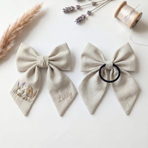 May include: Two beige linen hair bows. The left bow features embroidered wildflowers and the name Lilly in cursive script. The right bow has a black elastic hair tie attached to the centre. Both bows have long, pointed tails and a textured fabric.