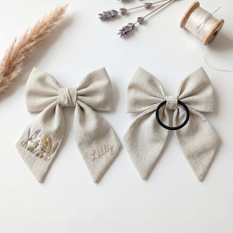 May include: Two beige linen hair bows. The left bow features embroidered wildflowers and the name Lilly in cursive script. The right bow has a black elastic hair tie attached to the centre. Both bows have long, pointed tails and a textured fabric.