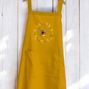 Puede incluir: Un delantal amarillo con un bolsillo grande y una abeja bordada en un círculo de flores amarillas.