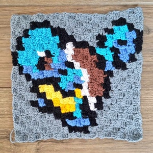 May include: A crocheted square with a pixelated design featuring a blue, brown, yellow, white, and black pattern. The square is made with gray yarn.