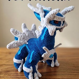 May include: A crocheted figure of a blue and white mythical creature, with the text "PDF PATTERN" at the top. The creature has a detailed design with spikes and a red eye. The figure is on a wooden surface.