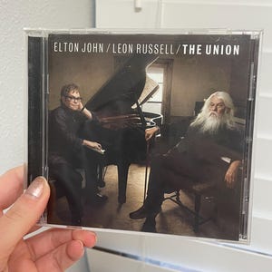 May include: CD case featuring Elton John and Leon Russell, with the title "The Union." The cover art shows the musicians seated near a grand piano. The image has a warm, sepia tone, suggesting a vintage or classic feel.