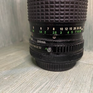 Puede incluir: Un objetivo de c&aacute;mara negro con una distancia focal de 200 mm y un anillo de apertura verde. El anillo de apertura tiene marcas para f/5.6, f/8, f/11, f/16, f/22 y f/32. El objetivo tambi&eacute;n tiene una escala de profundidad de campo verde con marcas para 1,5, 1,8, 2, 2,5, 3, 4, 5, 7, 10 y 20 metros.