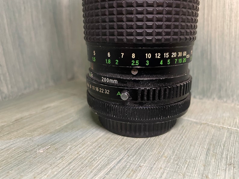 Puede incluir: Un objetivo de c&aacute;mara negro con una distancia focal de 200 mm y un anillo de apertura verde. El anillo de apertura tiene marcas para f/5.6, f/8, f/11, f/16, f/22 y f/32. El objetivo tambi&eacute;n tiene una escala de profundidad de campo verde con marcas para 1,5, 1,8, 2, 2,5, 3, 4, 5, 7, 10 y 20 metros.