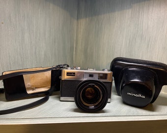 May include: A silver Minolta camera with a black lens and a black leather case. The camera has a silver body with black lettering and a black leather strap.