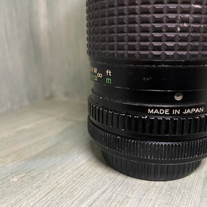 Puede incluir: Un objetivo de c&aacute;mara negro con textura acanalada y el texto "MADE IN JAPAN" impreso en el lateral. El objetivo tiene una escala verde y blanca con marcas para pies y metros.