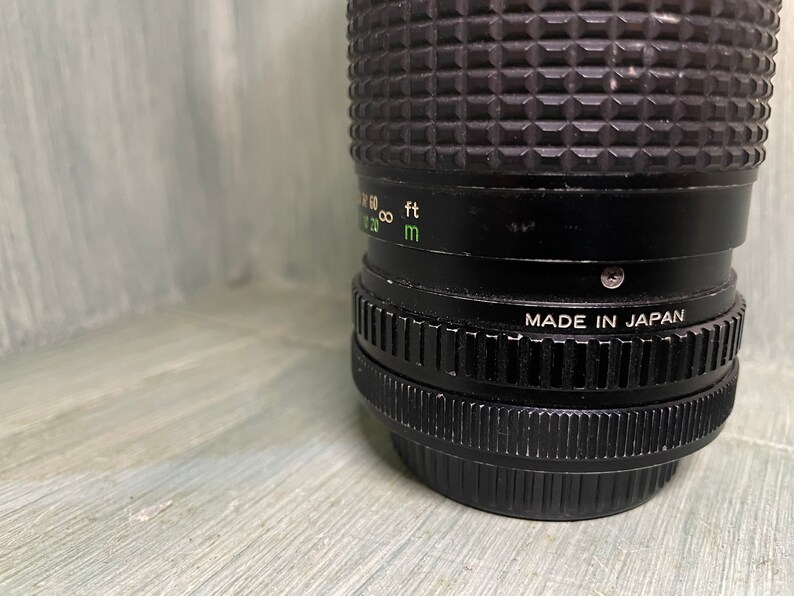 Puede incluir: Un objetivo de c&aacute;mara negro con textura acanalada y el texto "MADE IN JAPAN" impreso en el lateral. El objetivo tiene una escala verde y blanca con marcas para pies y metros.
