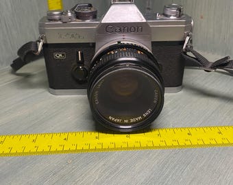Peut inclure: Un appareil photo Canon QL argenté avec un objectif Canon noir. L'objectif porte l'inscription "Lens Made in Japan".