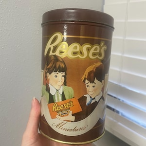 May include: A brown metal tin with a gold lid. The tin has a picture of two children holding a box of Reese's Peanut Butter Cups Miniatures. The text on the tin reads "Reese's Miniatures".