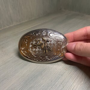 May include: Silver oval belt buckle with a gold-toned design of a cowboy riding a horse. The buckle has an ornate border.