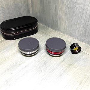 May include: Three gray and black camera lens filters in a black leather case. The filters are round and have a red and white label.