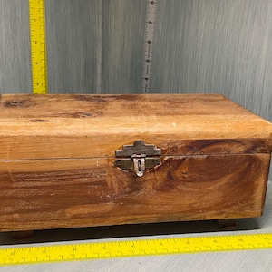 May include: A wooden box with a hinged lid and a metal latch. The box is made of light brown wood with a natural grain pattern. The box has a rustic appearance and is likely handmade.