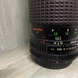 Puede incluir: Un objetivo de c&aacute;mara negro con un anillo y marcas plateadas. El objetivo tiene un ajuste macro y una distancia focal de 200 mm. Los ajustes de apertura son 4,556, 8, 11, 16, 22 y 32.
