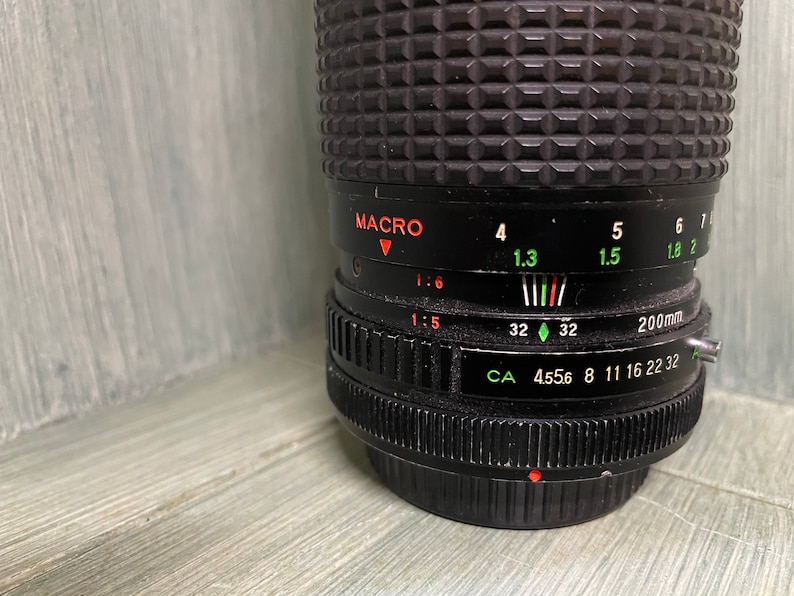 Puede incluir: Un objetivo de c&aacute;mara negro con un anillo y marcas plateadas. El objetivo tiene un ajuste macro y una distancia focal de 200 mm. Los ajustes de apertura son 4,556, 8, 11, 16, 22 y 32.